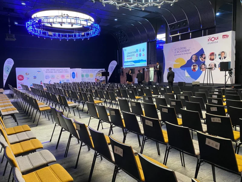 Vue d’ensemble de la salle avant la convention TGV INOUI Atlantique, avec scène, mur d’image, stands et drapeaux imprimés.
